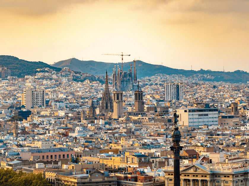 Panoramic view of Barcelona cityscape featuring the Sagrada Familia basilica and surrounding urban landscape with a backdrop of hills and a golden sky.