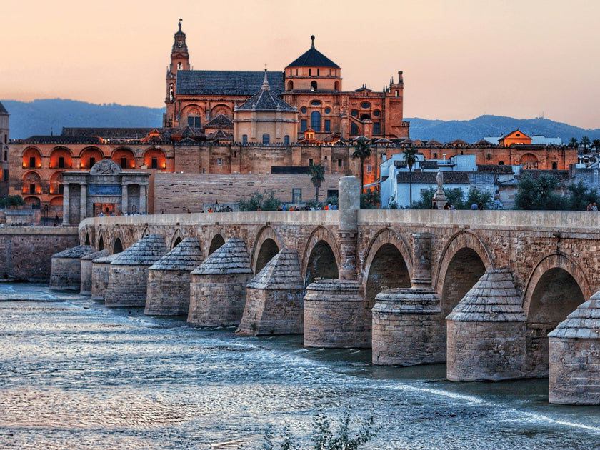 Roman Bridge in Córdoba with the iconic Mezquita-Catedral in the background during sunset, showcasing the historic architecture and serene river view in Spain.