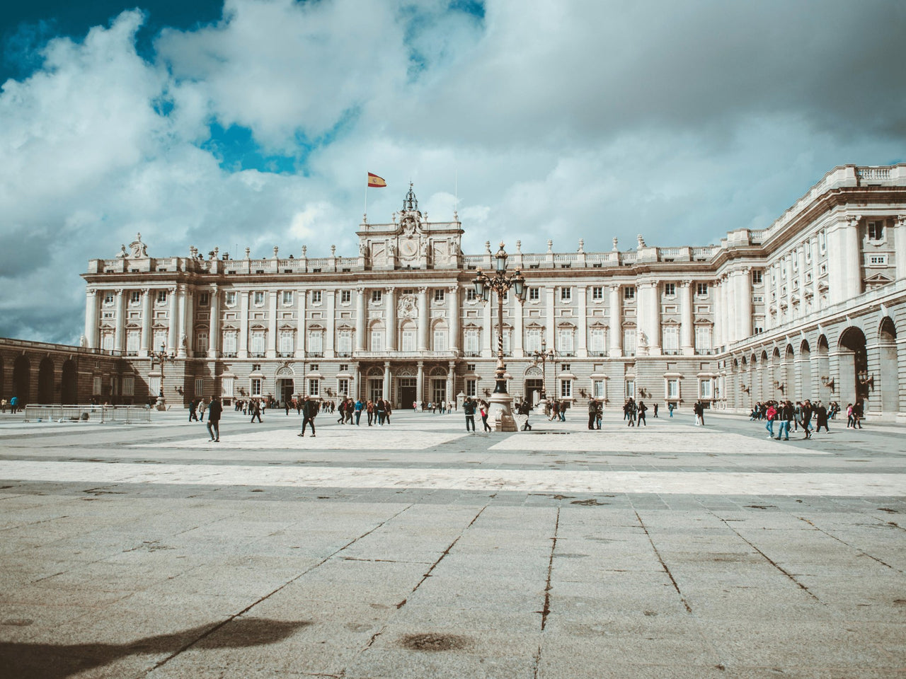 he Royal Palace of Madrid, Spain, featuring neoclassical architecture with an expansive courtyard and people walking under a partly cloudy sky.