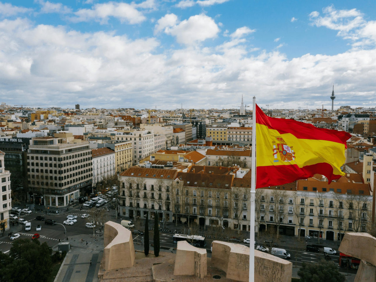 Panoramic view of Madrid, Spain, with the Spanish flag waving in the foreground against a backdrop of city buildings and a partly cloudy sky.