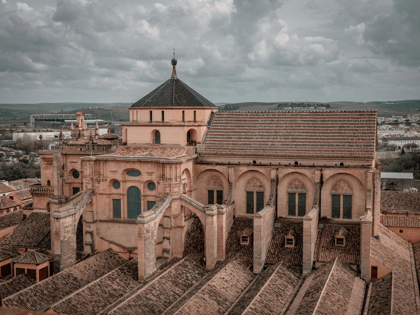 Rooftop view of the Mezquita-Catedral (Mosque-Cathedral) of Córdoba, Spain, with intricate architectural details under a cloudy sky and a scenic background.